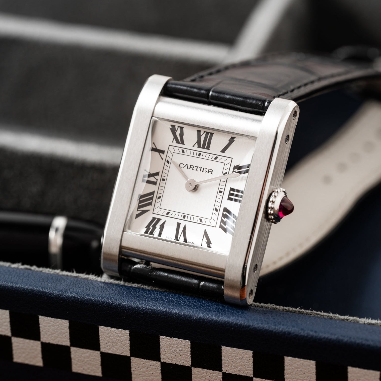 A Cartier Tank Normale wristwatch with a square face, Roman numerals, and a black leather strap on a patterned surface.