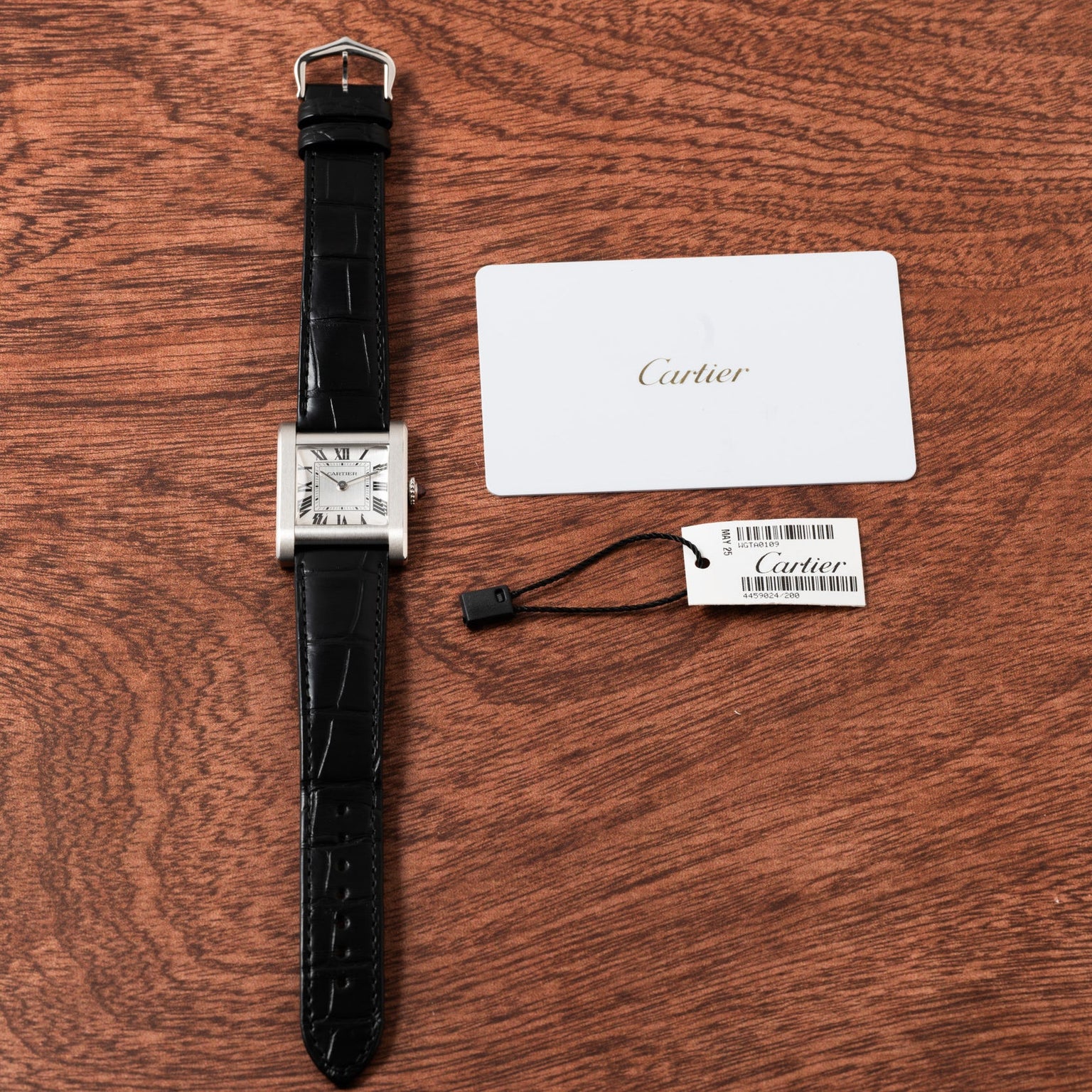 A Cartier Tank Normale wristwatch with a black strap sits next to a Cartier tag and card on a wooden surface.
