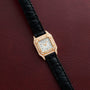 The Cartier Panthère gold watch features diamonds and a black leather strap, displayed on a textured dark red background.