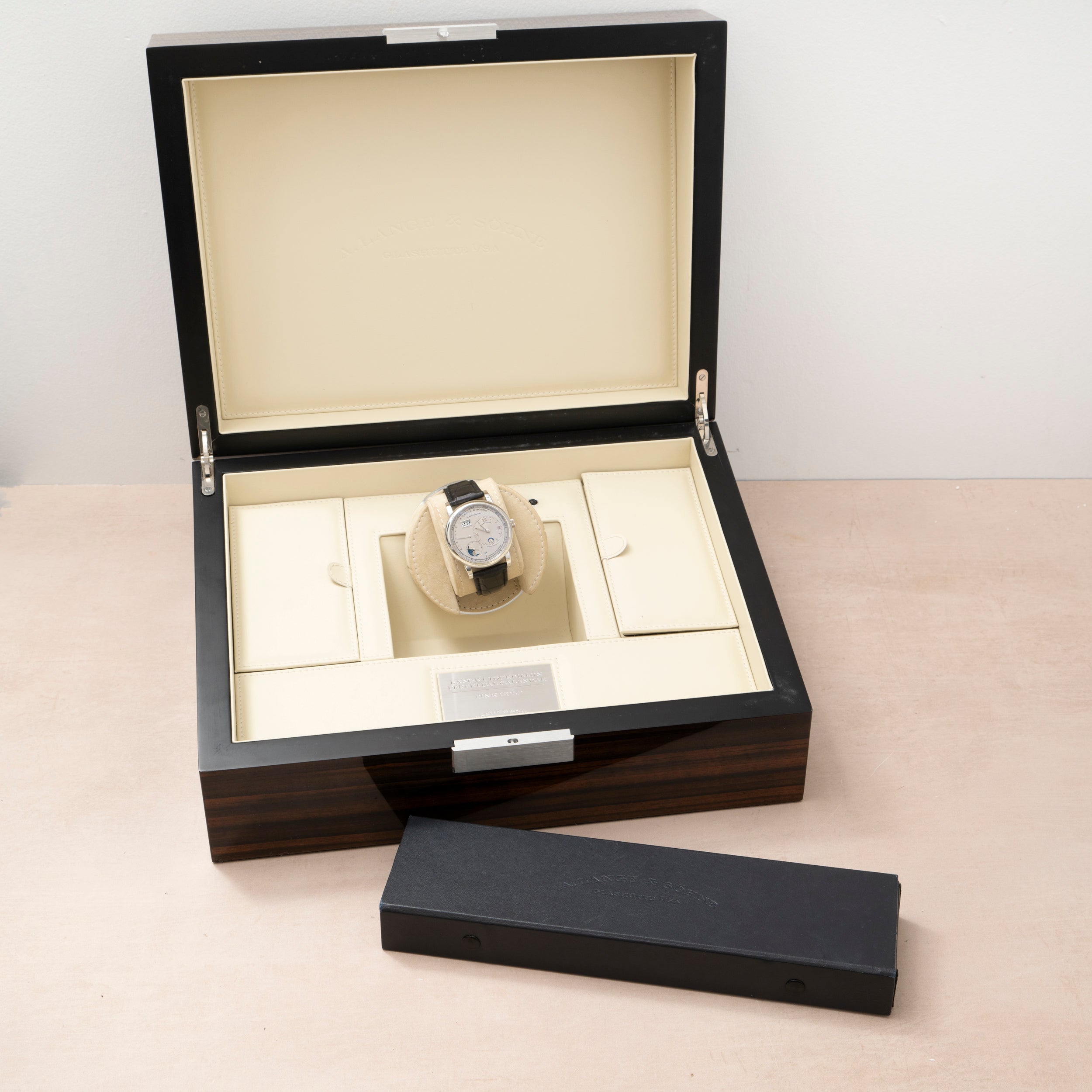 The Lange & Sohne Lange 1 Tourbillon Perpetual Calendar luxury wristwatch is displayed in an open wooden box with a long, closed black case in front.