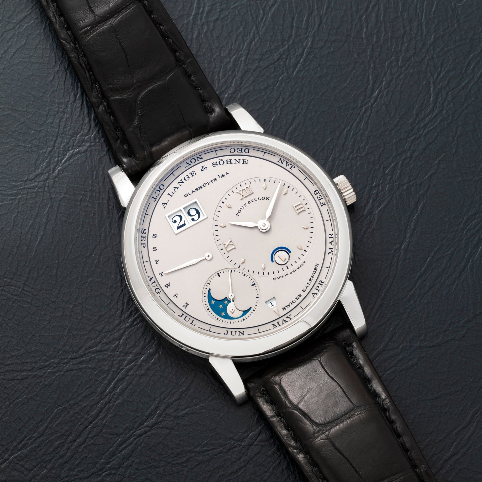 The Lange & Sohne Lange 1 Tourbillon Perpetual Calendar wristwatch features multiple dials and a black leather strap, displayed on a dark textured surface.