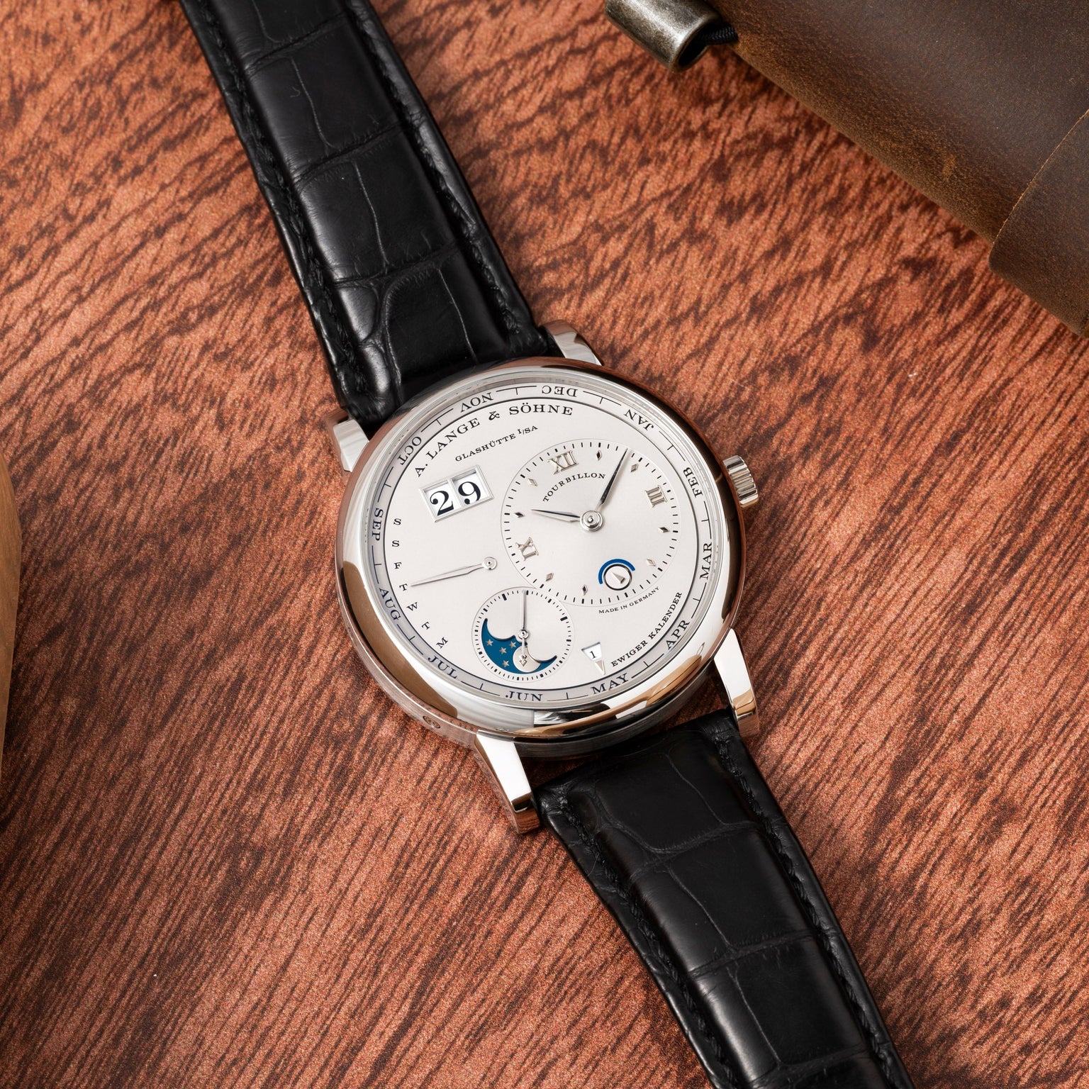 The Lange & Sohne Lange 1 Tourbillon Perpetual Calendar, featuring a black leather strap and silver dial, is elegantly displayed on a wooden surface.