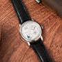 The Lange & Sohne Lange 1 Tourbillon Perpetual Calendar, featuring a black leather strap and silver dial, is elegantly displayed on a wooden surface.