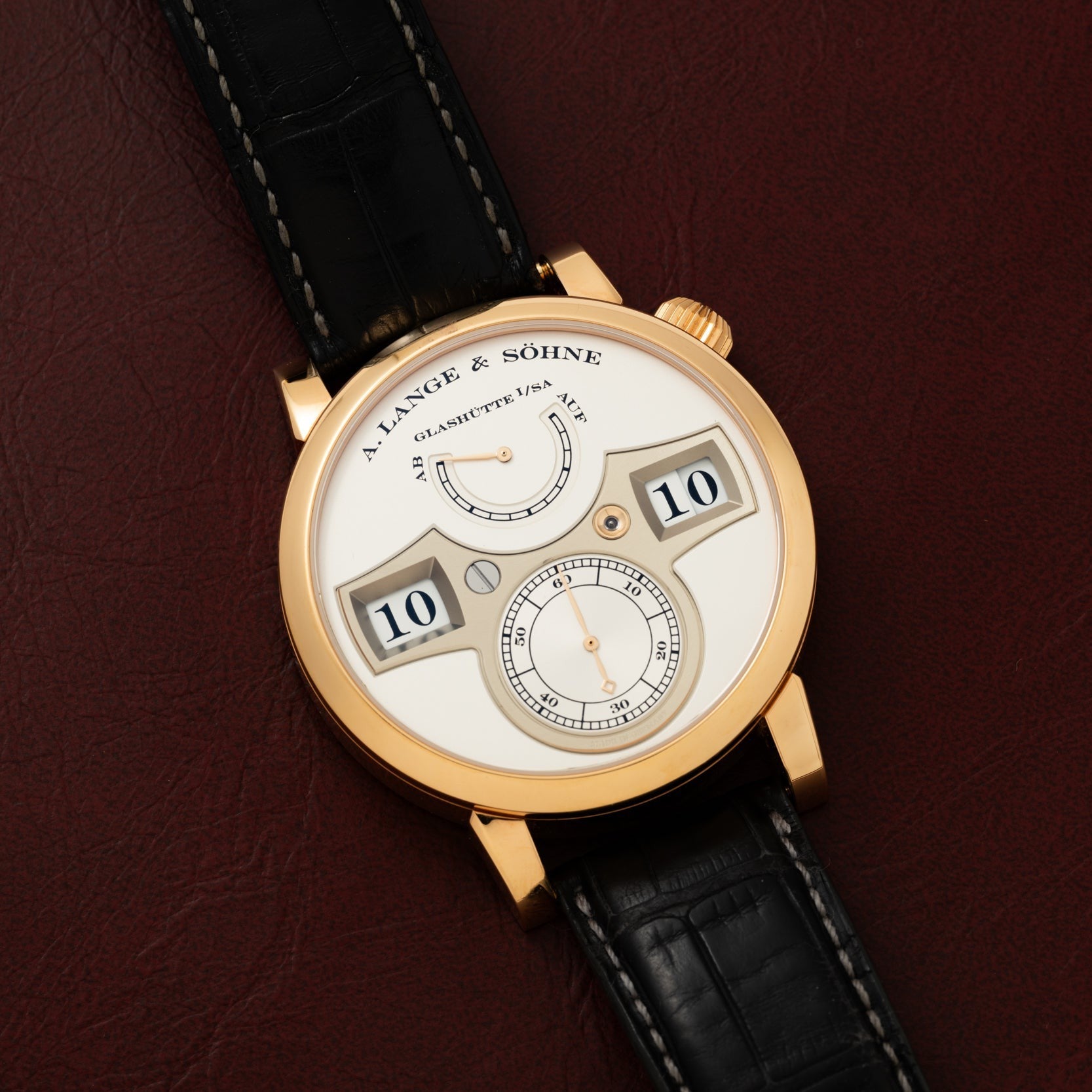 A Lange & Sohne Zeitwerk wristwatch featuring a gold case and black leather strap, displayed on a brown leather surface.