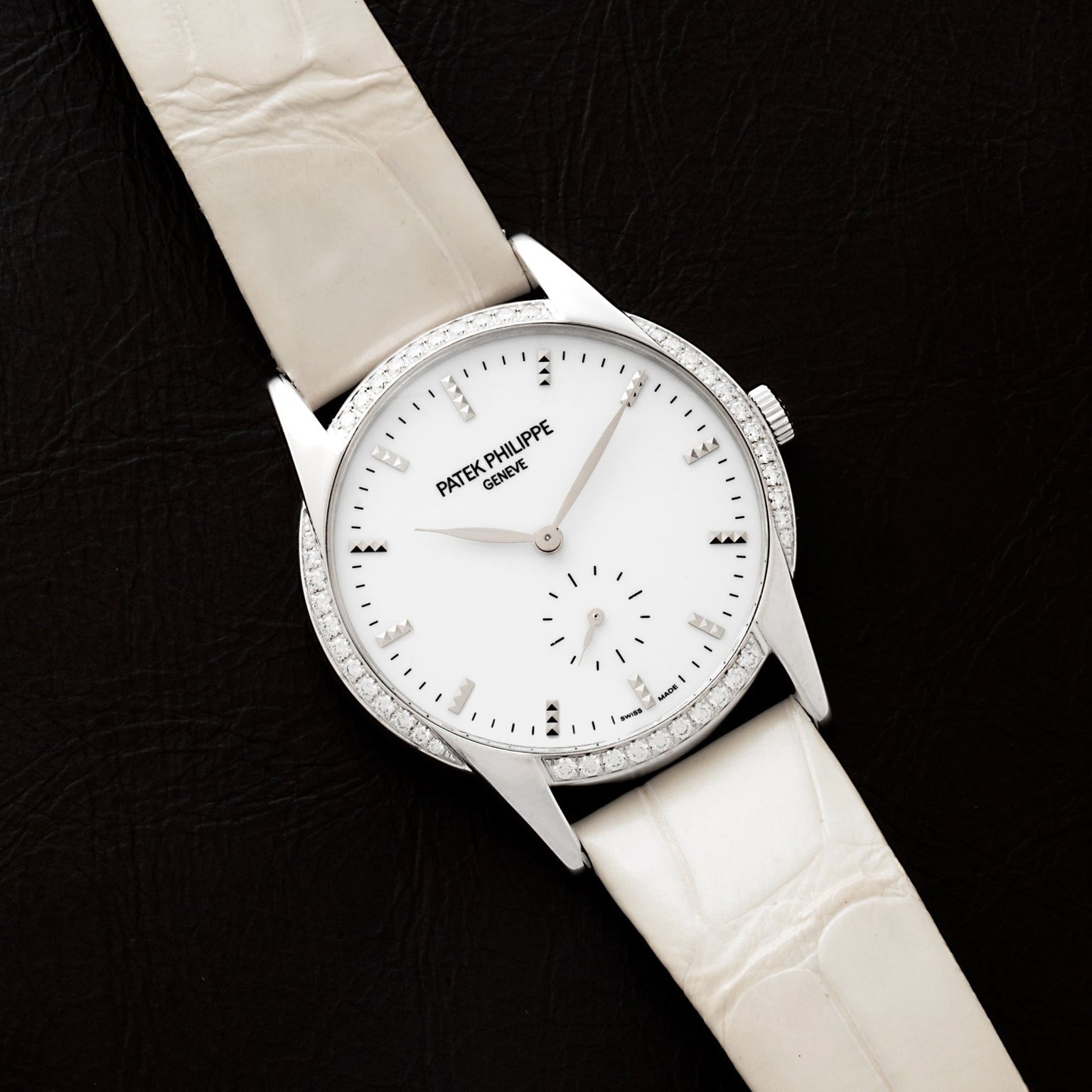 A Patek Philippe Calatrava wristwatch featuring a diamond bezel and white leather strap, displayed on a black surface.