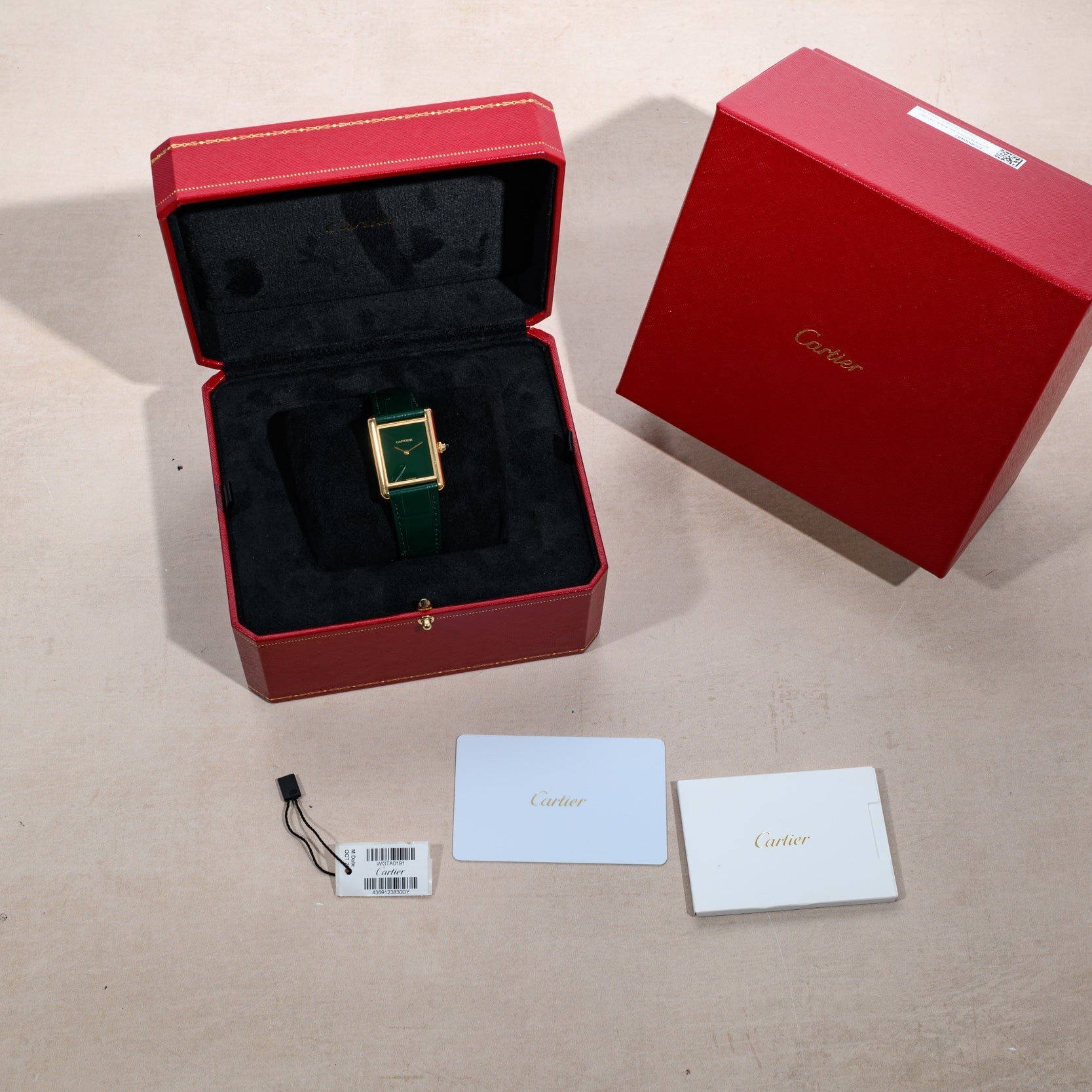 A Cartier Tank Louis watch in a red box, with tags, warranty card, and booklet arranged on a beige surface.