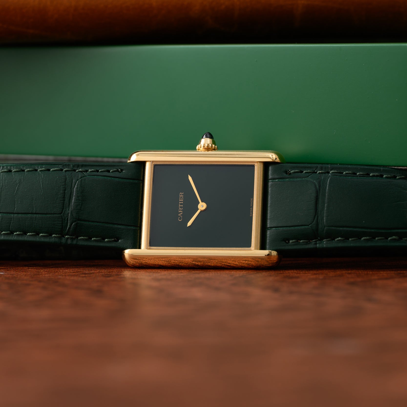 A Cartier Tank Louis watch with a black dial and green leather strap sits on a wooden surface.