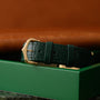 The Cartier Tank Louis green leather watch strap with a gold buckle rests on a matching green box.