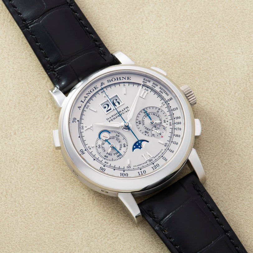 A luxurious Lange & Sohne Datograph Perpetual Calendar wristwatch with a black leather strap and multiple dials, displayed on a beige surface.
