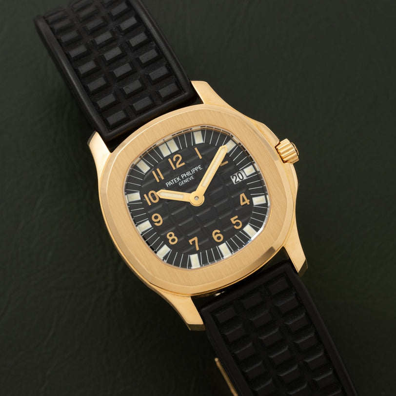 The Patek Philippe Aquanaut features a black textured strap and black dial, showcased on a dark surface.