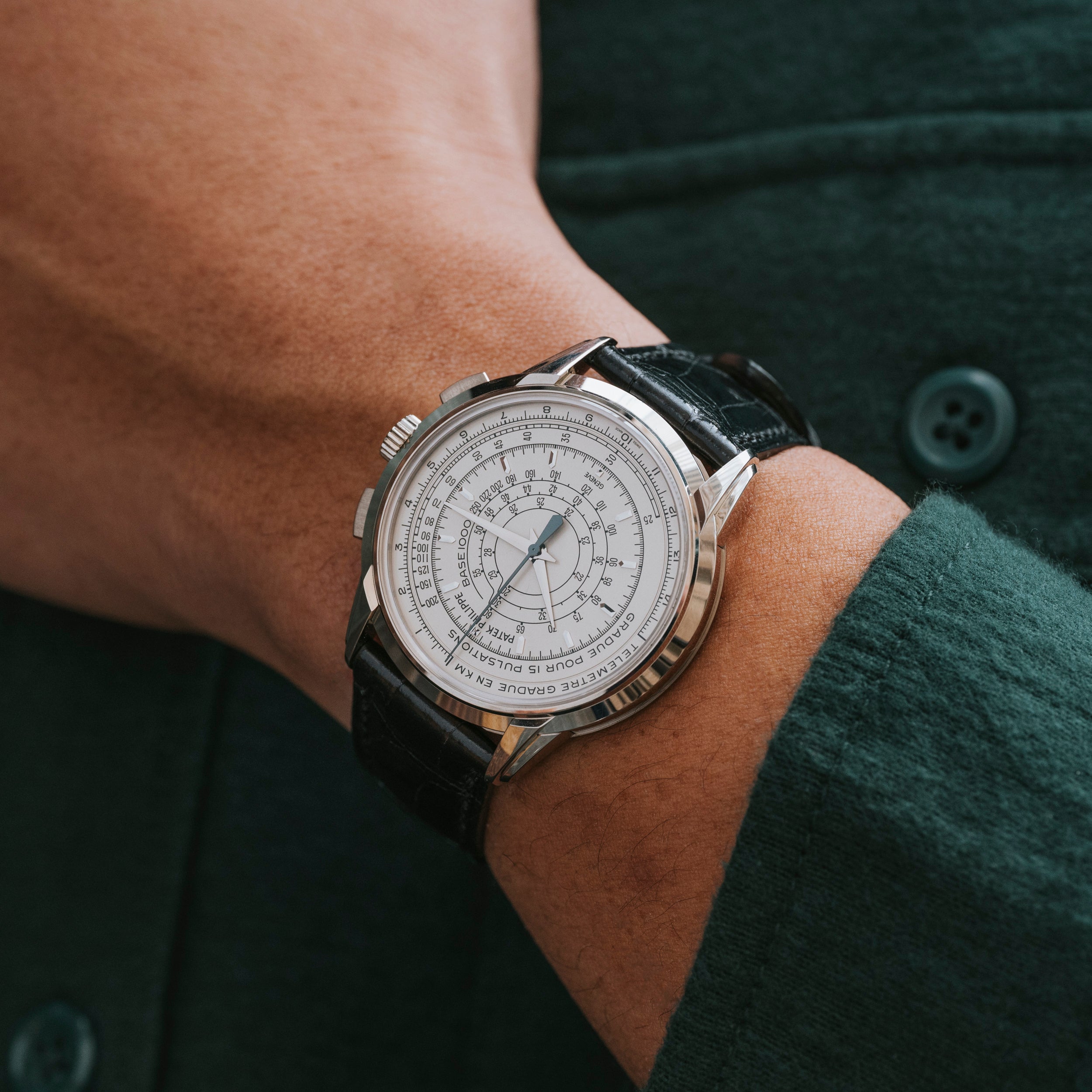 A person wearing the Patek Philippe Multi-Scale Chronograph, a luxury silver watch with a white dial and black leather strap, dressed in a green shirt.