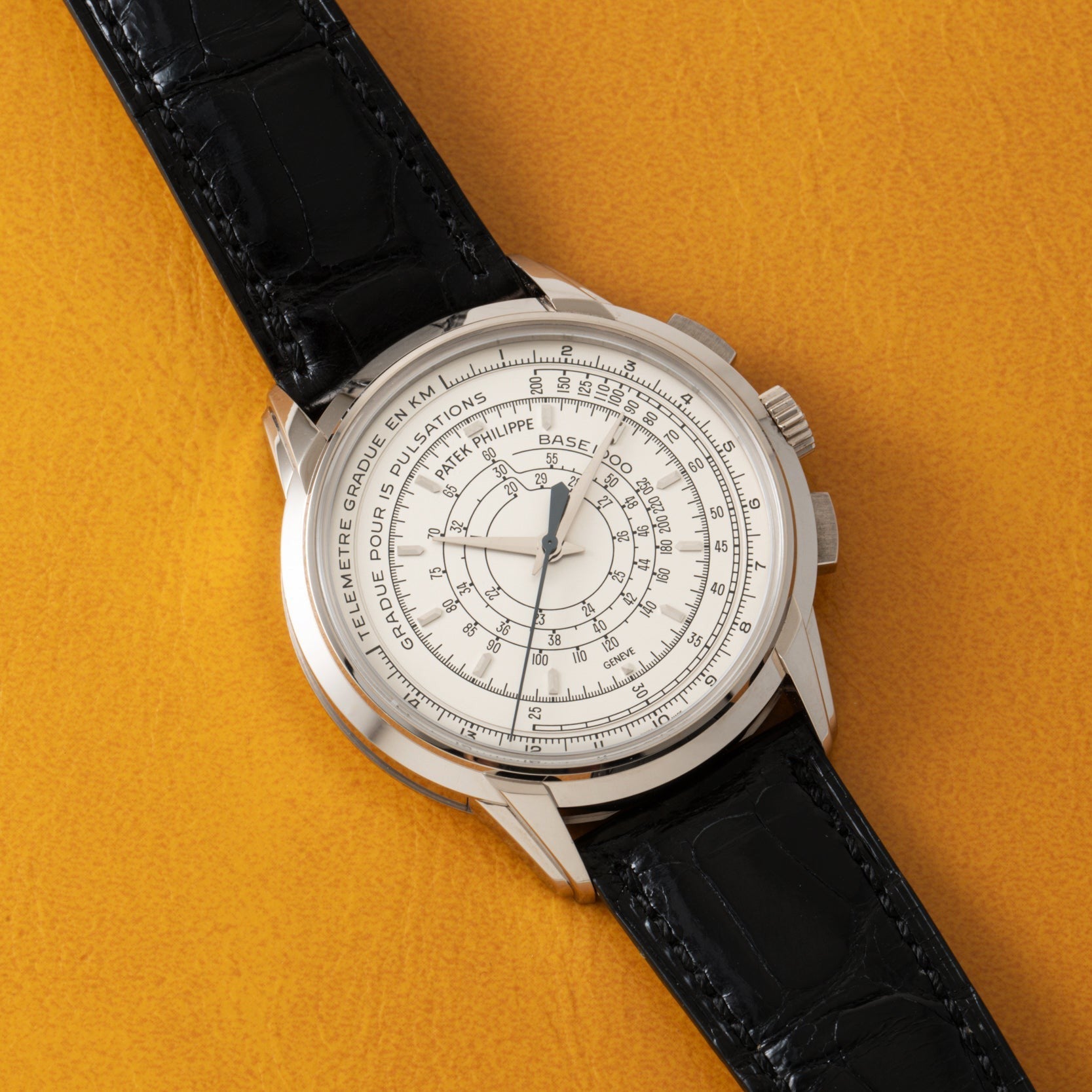 The Patek Philippe Multi-Scale Chronograph, featuring a black leather strap and an intricate white dial, is displayed on a yellow textured surface.