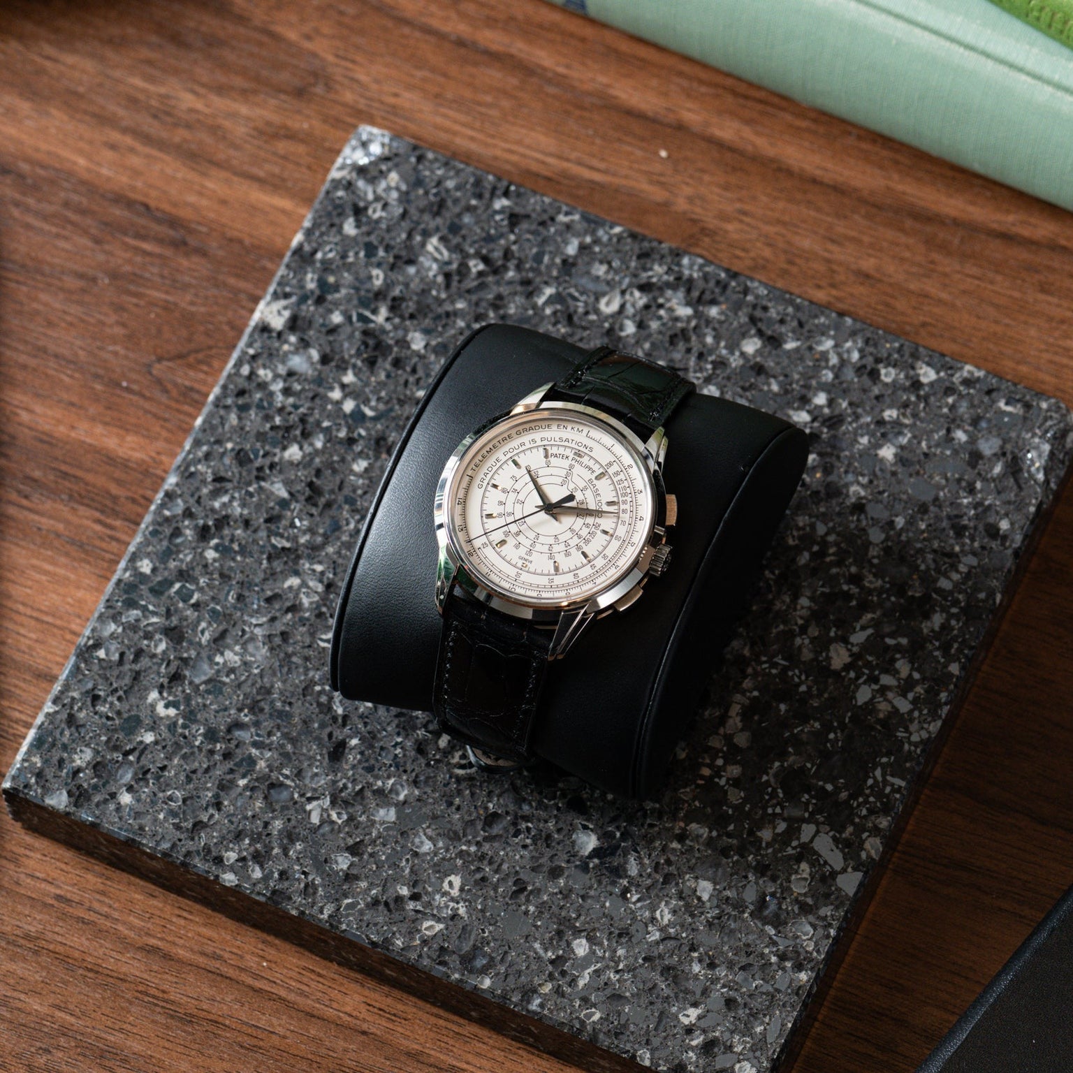 The Patek Philippe Multi-Scale Chronograph, featuring a silver case and black leather band, is displayed on a black stand atop a speckled stone surface.