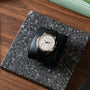 The Patek Philippe Multi-Scale Chronograph, featuring a silver case and black leather band, is displayed on a black stand atop a speckled stone surface.