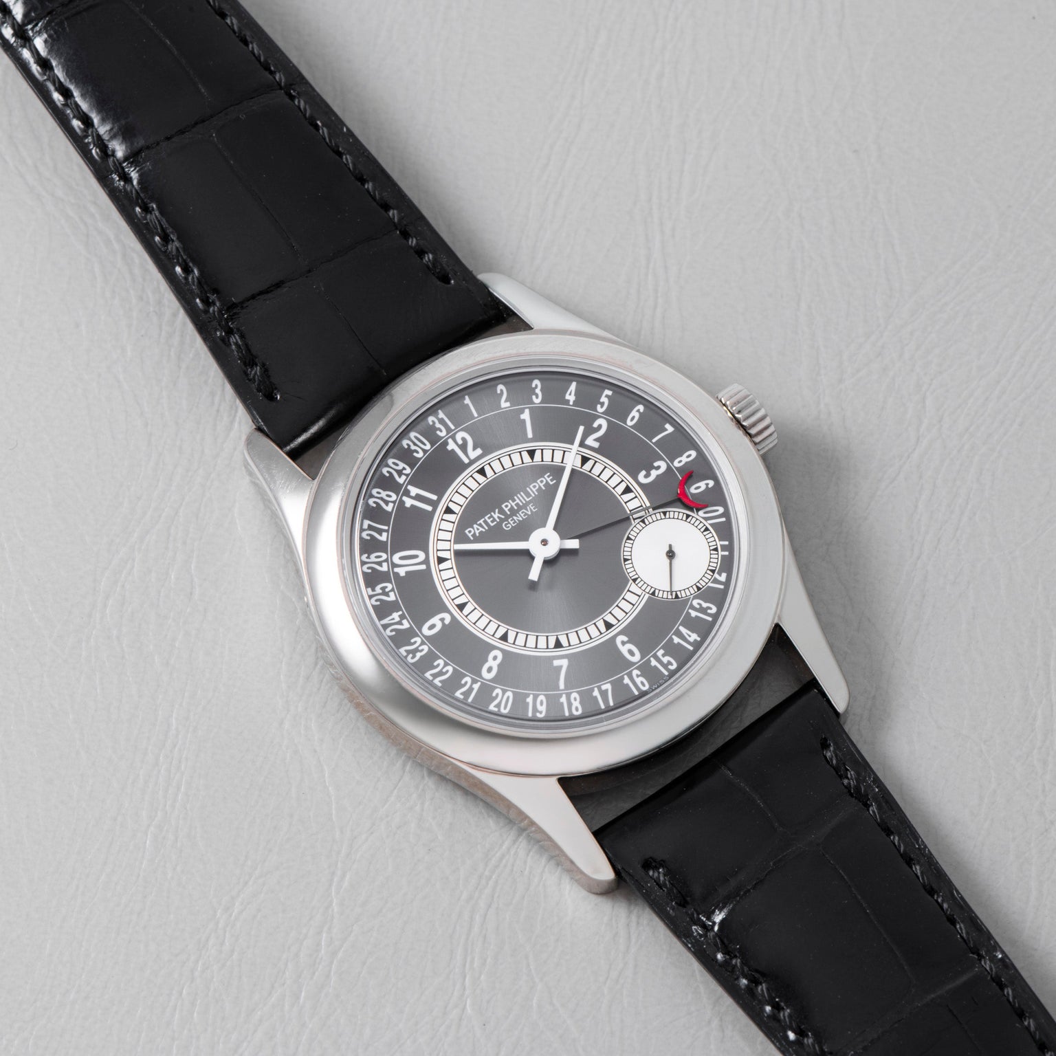 A Patek Philippe Calatrava wristwatch with a black leather strap rests on a light gray surface.