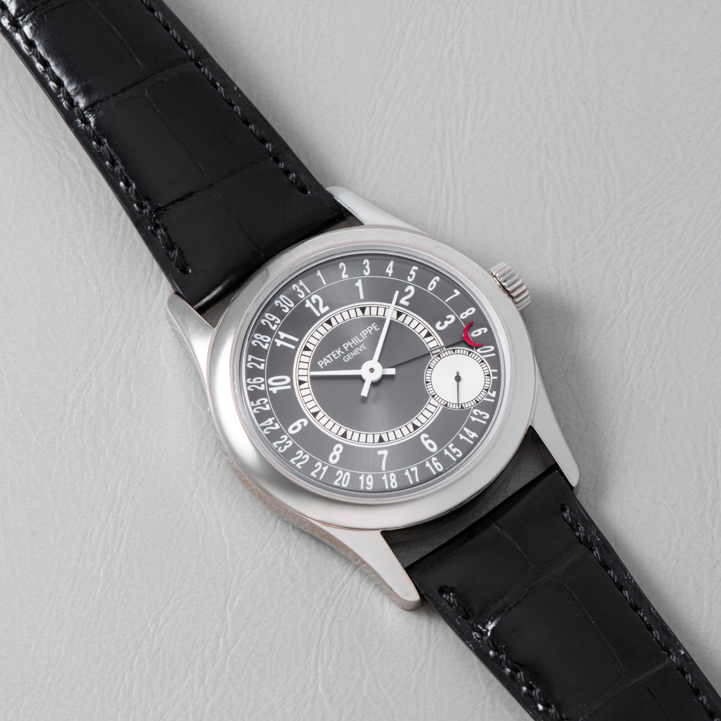 A Patek Philippe Calatrava wristwatch with a black leather strap rests on a light gray surface.