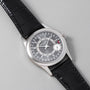 A Patek Philippe Calatrava wristwatch with a black leather strap rests on a light gray surface.