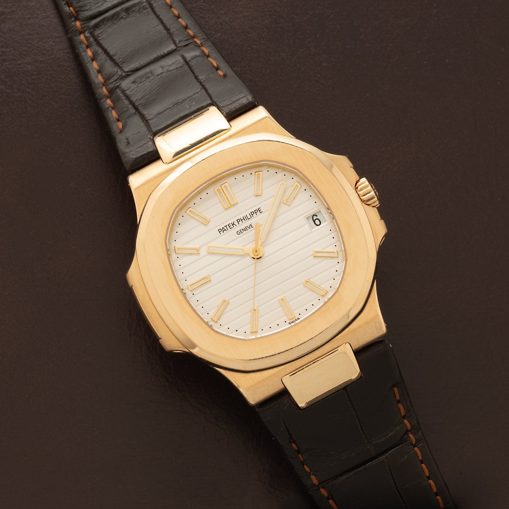 A Patek Philippe Nautilus wristwatch with a white dial and black leather strap is shown on a brown surface.
