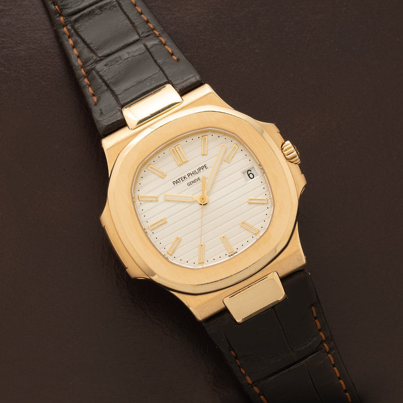 A Patek Philippe Nautilus wristwatch with a white dial and black leather strap is shown on a brown surface.