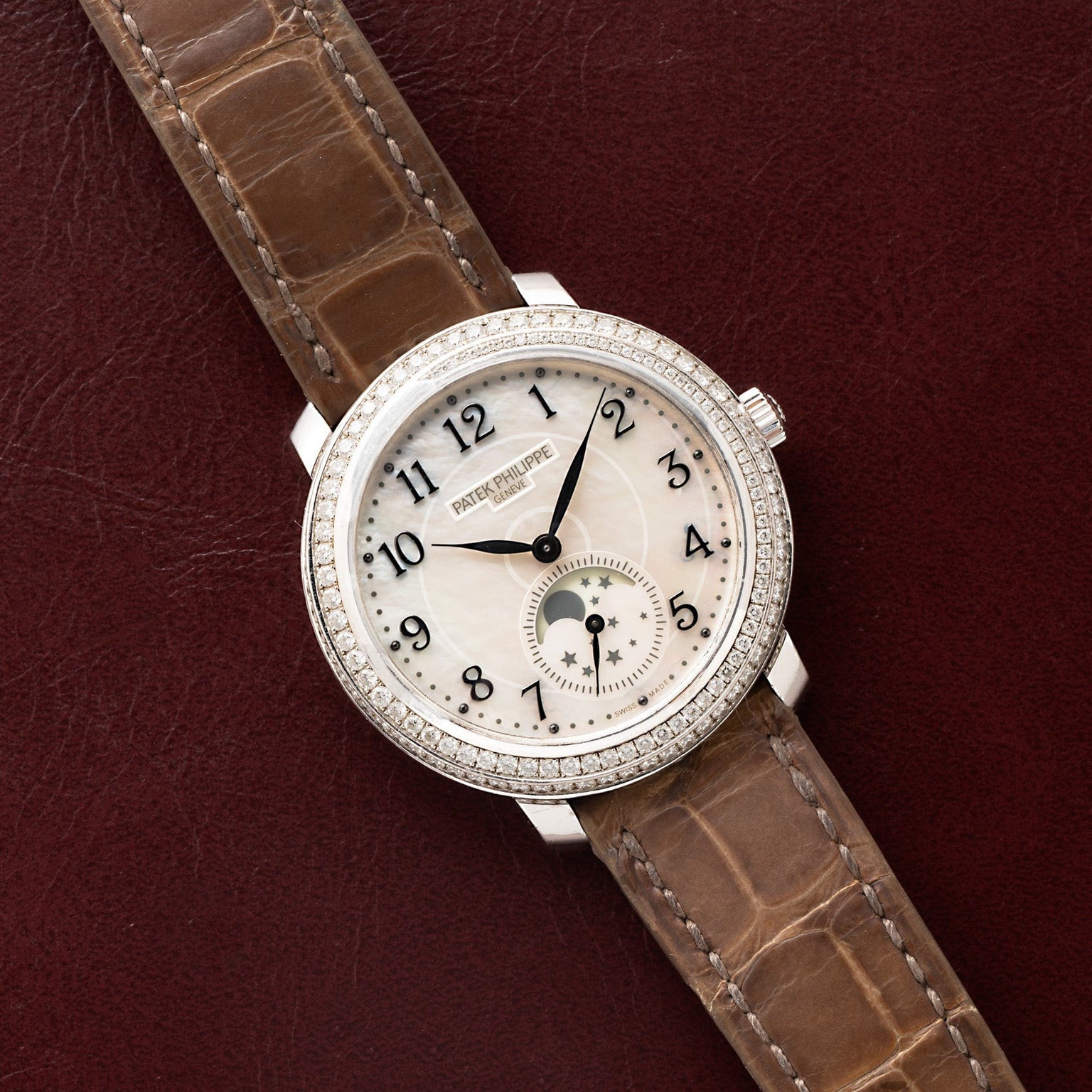 The Patek Philippe Ladies Complication watch, featuring a brown leather strap, is displayed on a dark red surface.