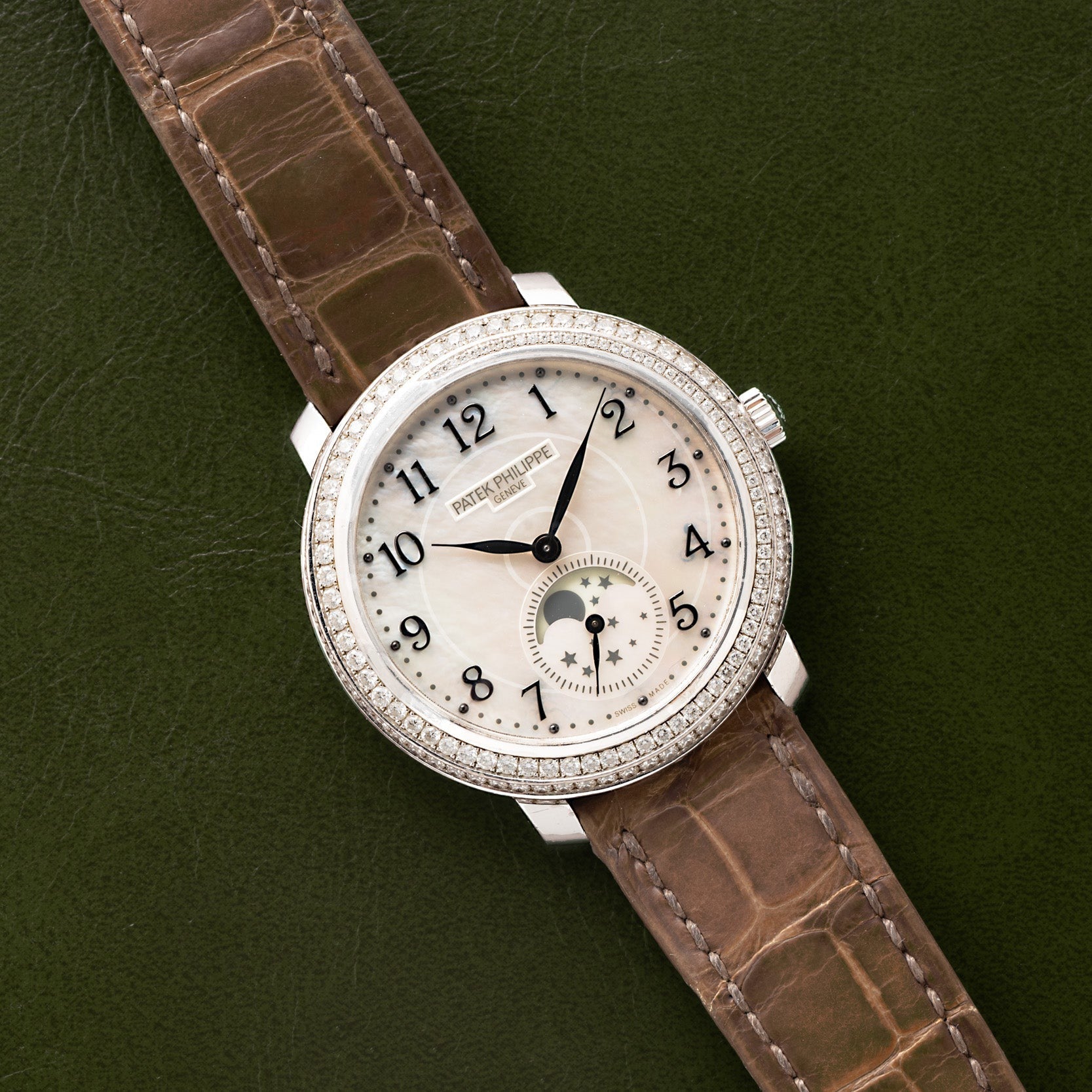 The Patek Philippe Ladies Complication features a brown strap, diamond bezel, and moonphase dial, displayed on a green surface.