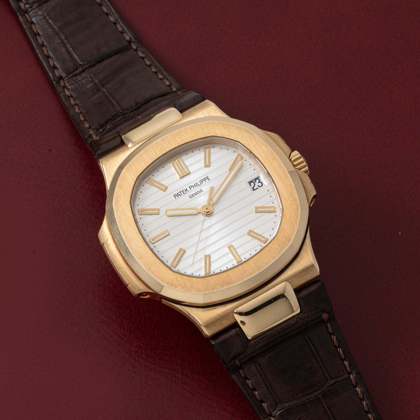 Patek Philippe Nautilus watch with white dial and brown leather strap on a dark red surface.