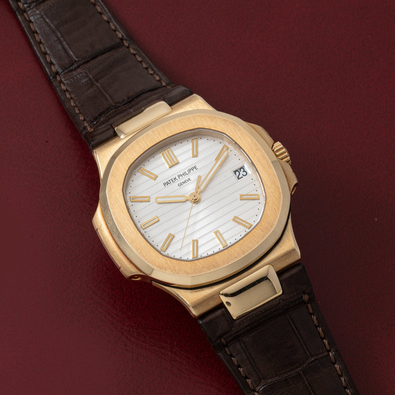 Patek Philippe Nautilus watch with white dial and brown leather strap on a dark red surface.