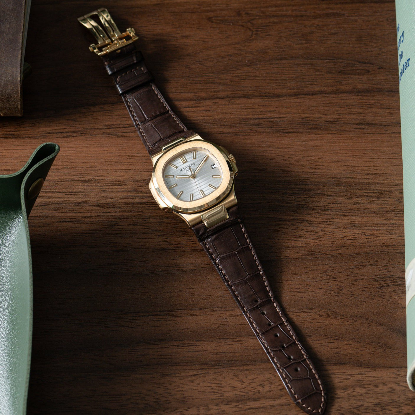 The Patek Philippe Nautilus gold wristwatch with a brown leather band rests on a wooden surface, partially surrounded by objects.