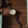 The Patek Philippe Nautilus gold wristwatch with a brown leather band rests on a wooden surface, partially surrounded by objects.
