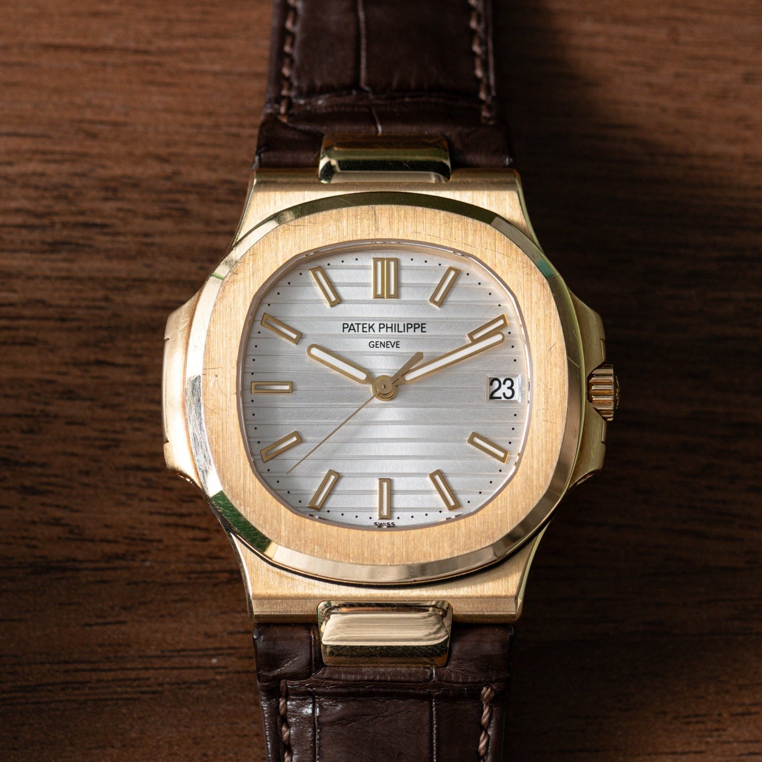 Patek Philippe Nautilus gold watch with a brown leather strap rests on a wooden surface, displaying the date as 23.