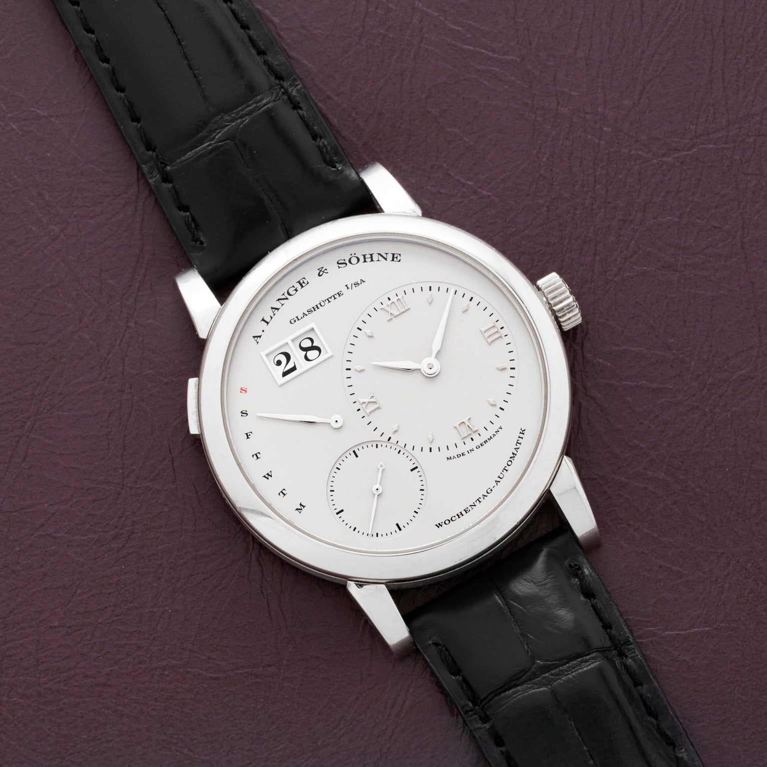A Lange & Sohne Lange 1 Daymatic wristwatch with a white dial and black leather strap displayed on a purple leather surface.