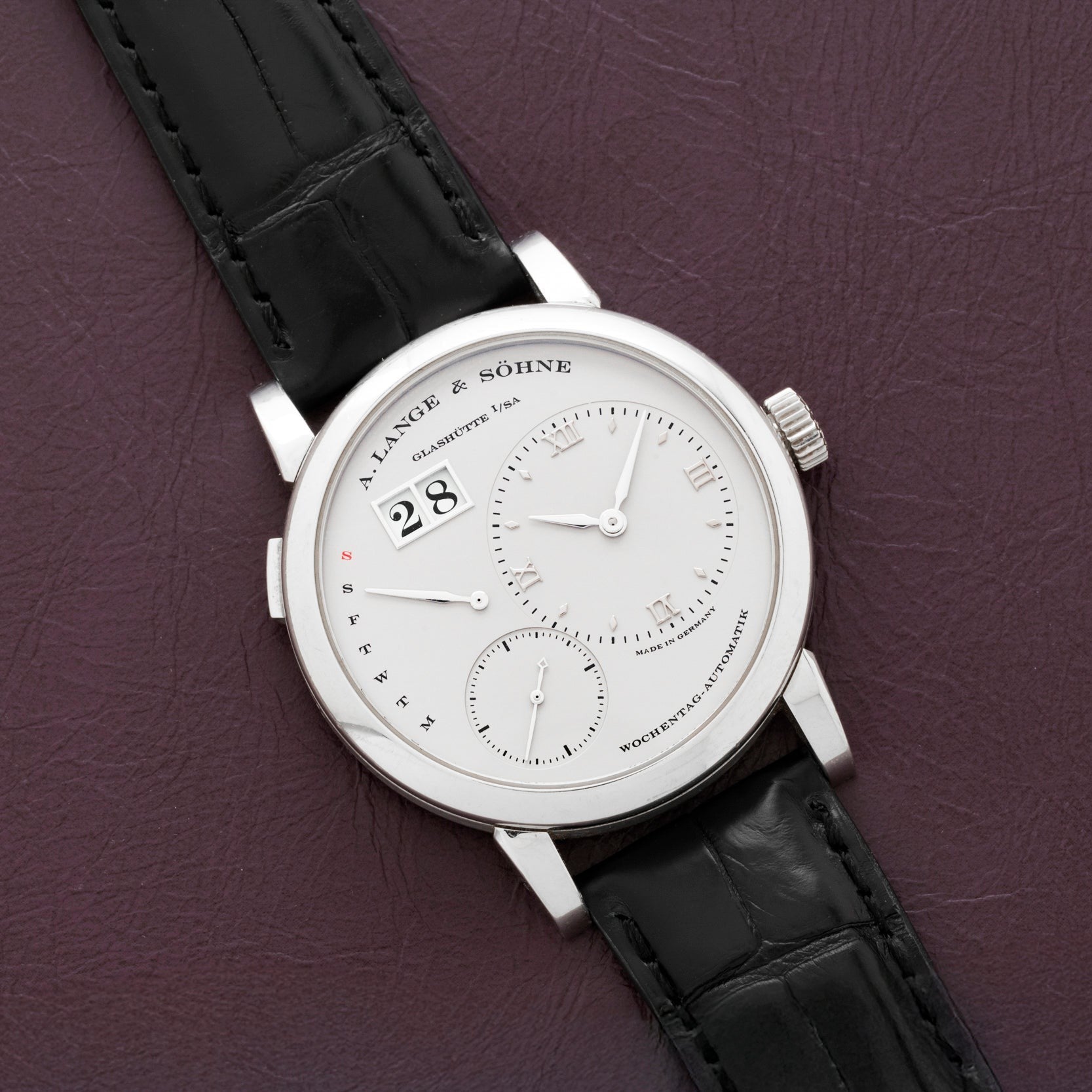 A Lange & Sohne Lange 1 Daymatic wristwatch with a white dial and black leather strap displayed on a purple leather surface.