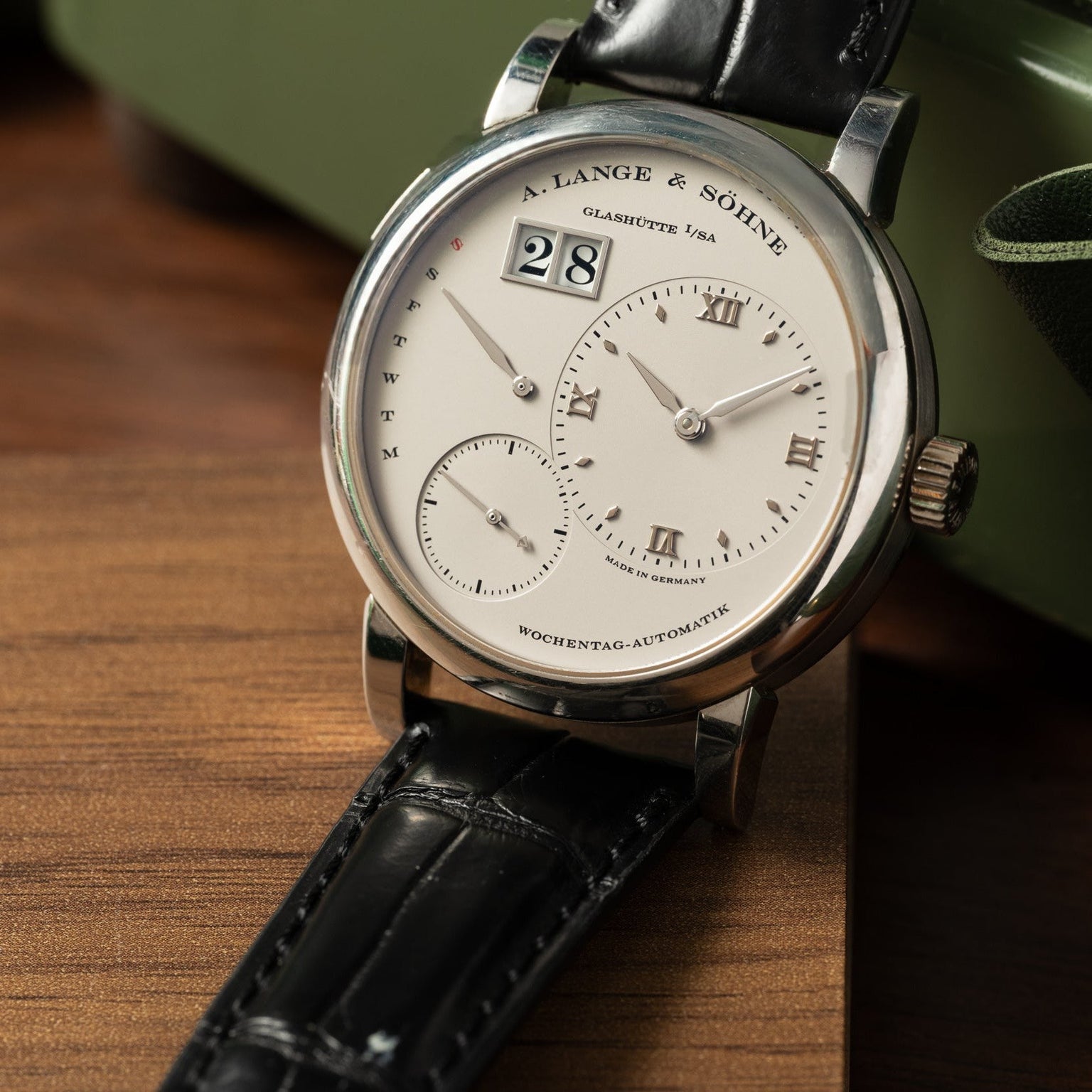A Lange & Sohne Lange 1 Daymatic watch with a silver dial and black leather strap displayed on a wooden surface.