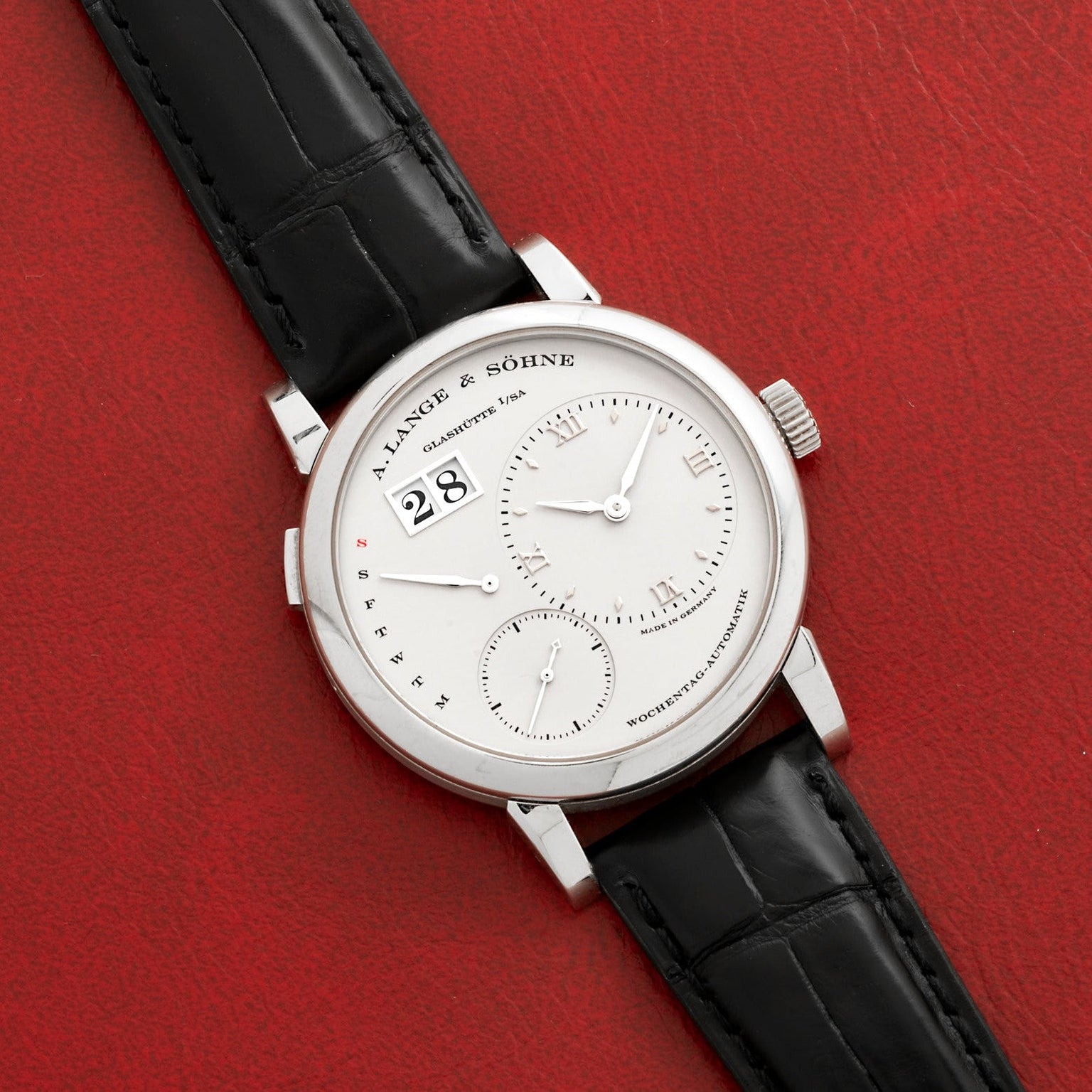 A Lange & Sohne Lange 1 Daymatic wristwatch with a black leather strap rests on a red textured surface.