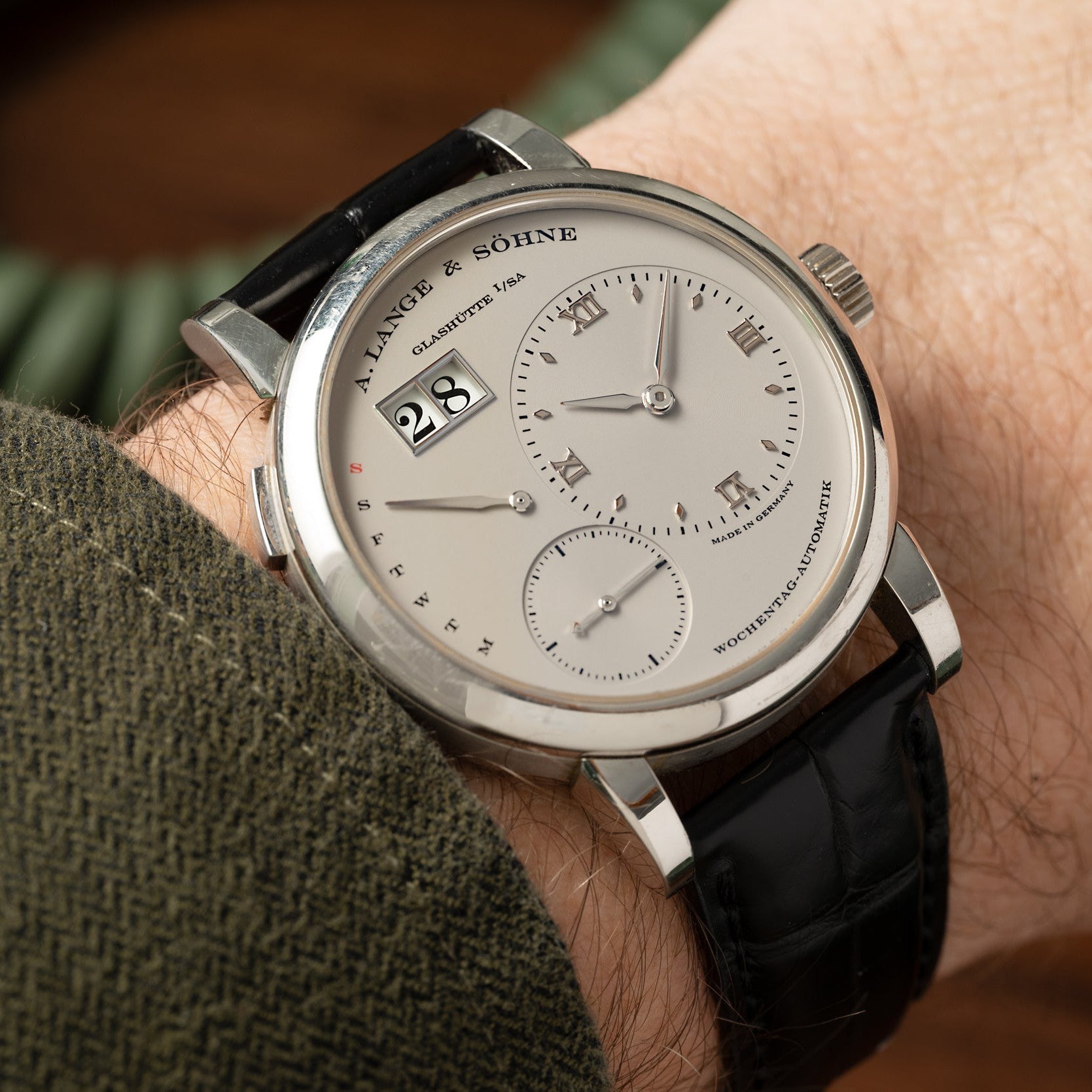 The Lange & Sohne Lange 1 Daymatic is a wristwatch with a silver dial, date display, and black leather strap, worn on the wrist.