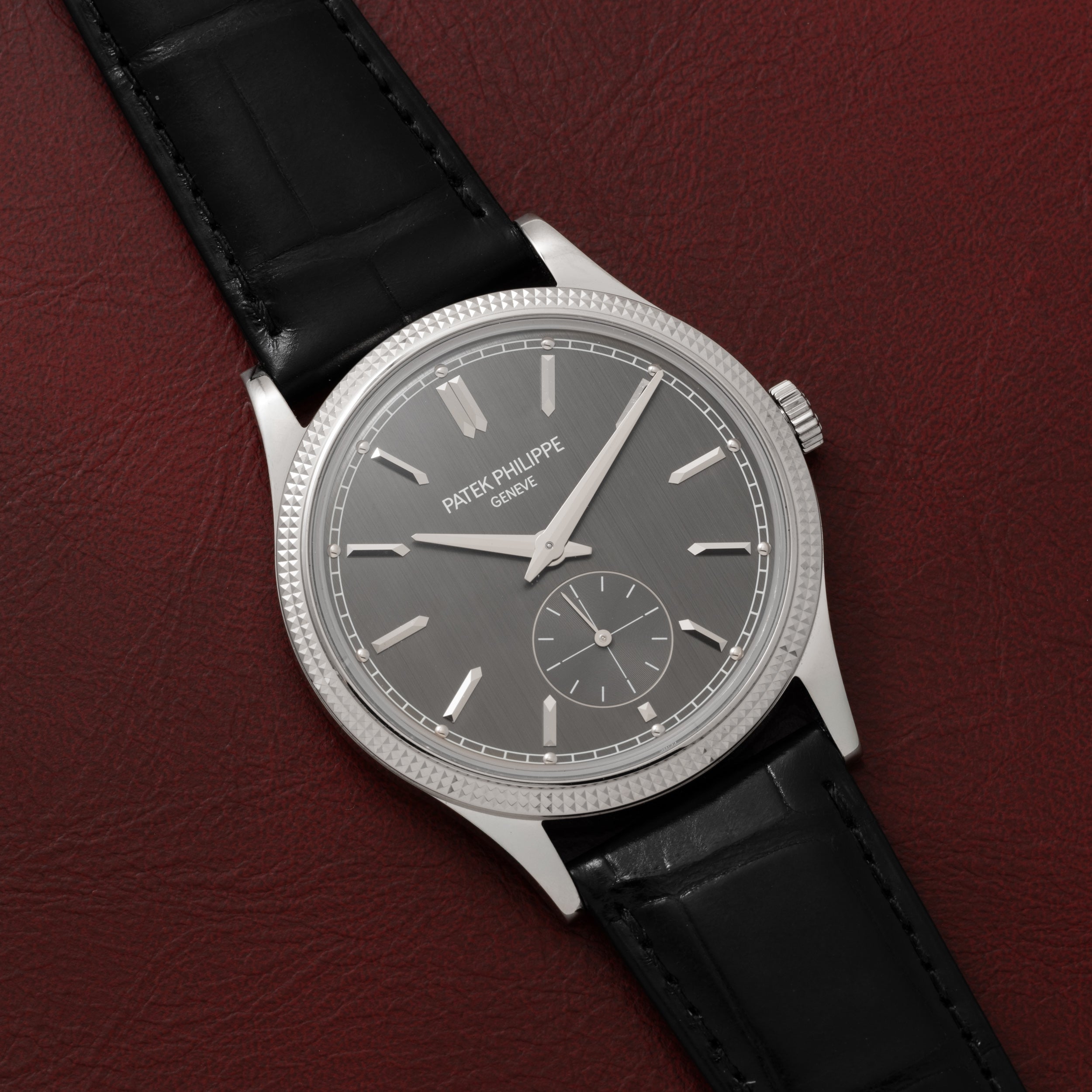 A Patek Philippe Calatrava with a silver case, gray dial, and black leather strap rests on a burgundy leather surface.
