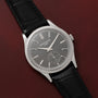 A Patek Philippe Calatrava with a silver case, gray dial, and black leather strap rests on a burgundy leather surface.