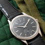 A close-up of the Patek Philippe Calatrava watch with a black leather strap on a green quilted fabric background.