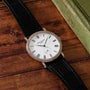 A Patek Philippe Calatrava wristwatch with a black leather strap rests on a wooden surface beside a green book.