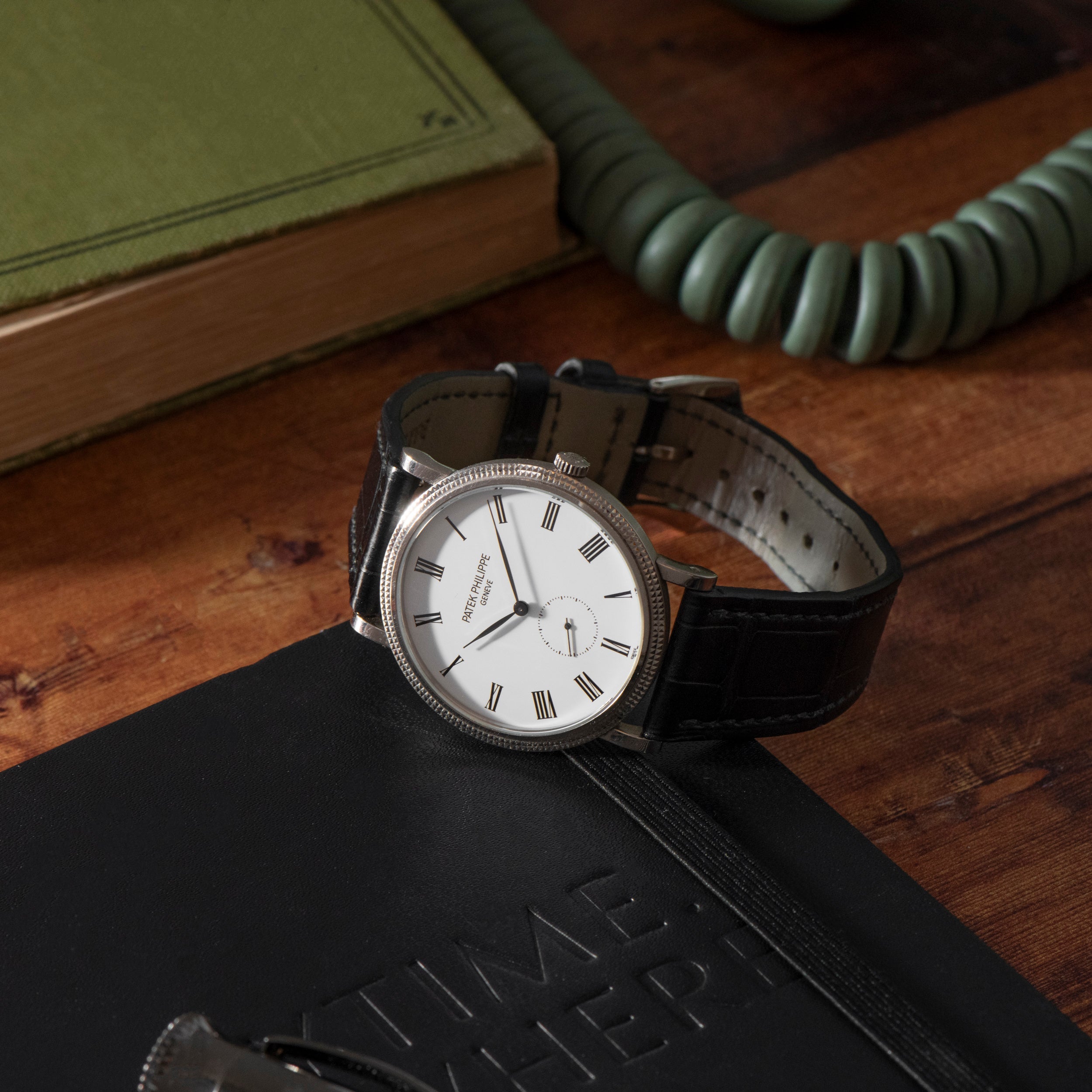A Patek Philippe Calatrava wristwatch with a black leather strap rests on a black notebook on a wooden desk.