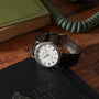 A Patek Philippe Calatrava wristwatch with a black leather strap rests on a black notebook on a wooden desk.