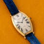 Cartier Tonneau XL watch with a blue leather strap, displayed on a yellow surface, featuring Roman numerals on its face.