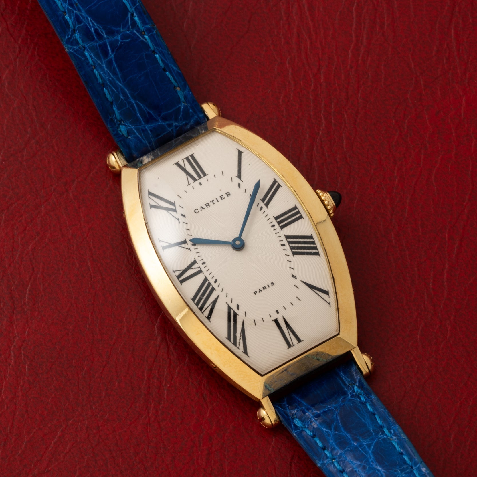 Cartier Tonneau XL wristwatch with Roman numerals and blue leather strap, displayed on a red textured surface.