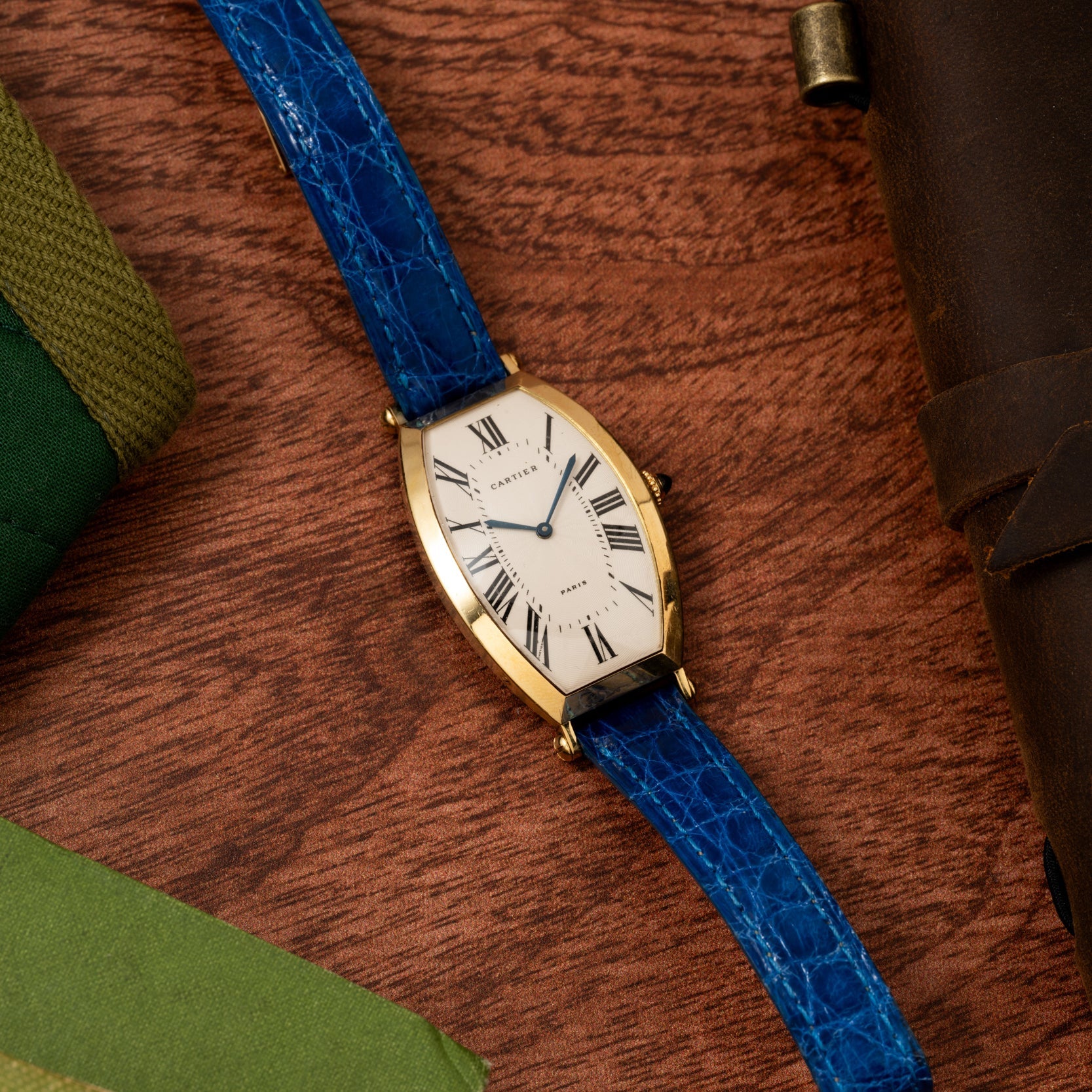 The Cartier Tonneau XL gold watch with a blue leather band rests on a wooden surface, surrounded by green and brown items.
