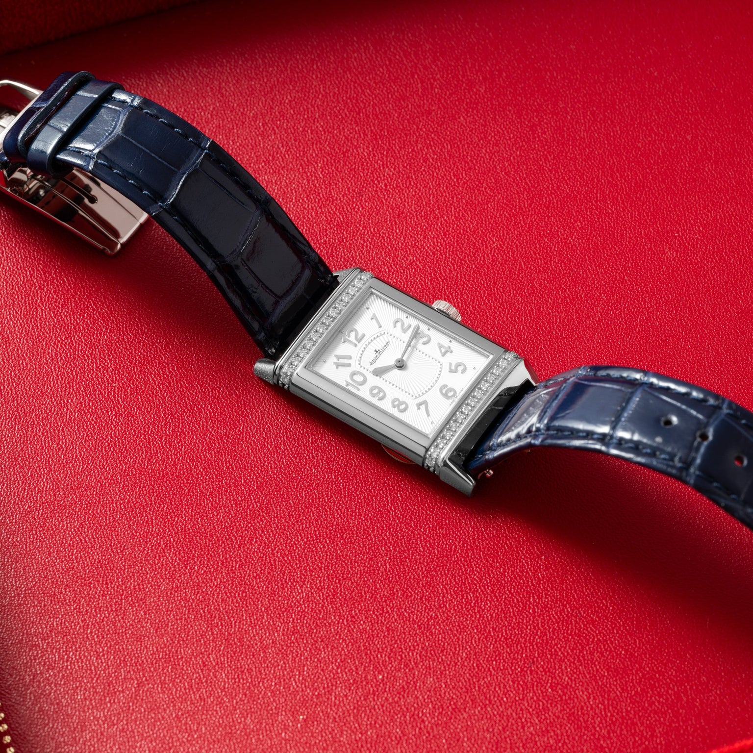 The Jaeger-LeCoultre Reverso Classique Ladies, a rectangular silver watch by Jaeger-LeCoultre with a blue leather strap, displayed on a red leather surface.