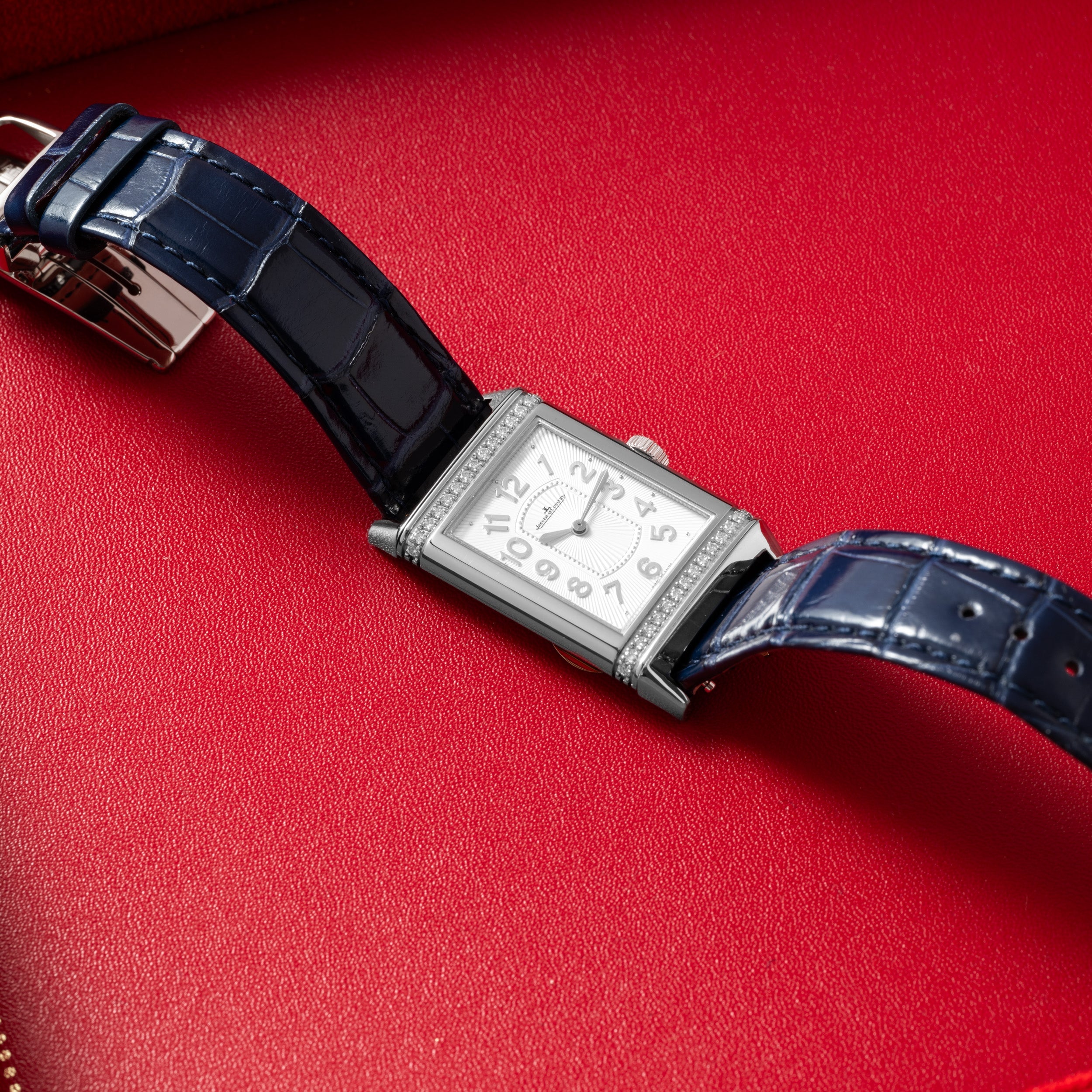 The Jaeger-LeCoultre Reverso Classique Ladies, a rectangular silver watch by Jaeger-LeCoultre with a blue leather strap, displayed on a red leather surface.
