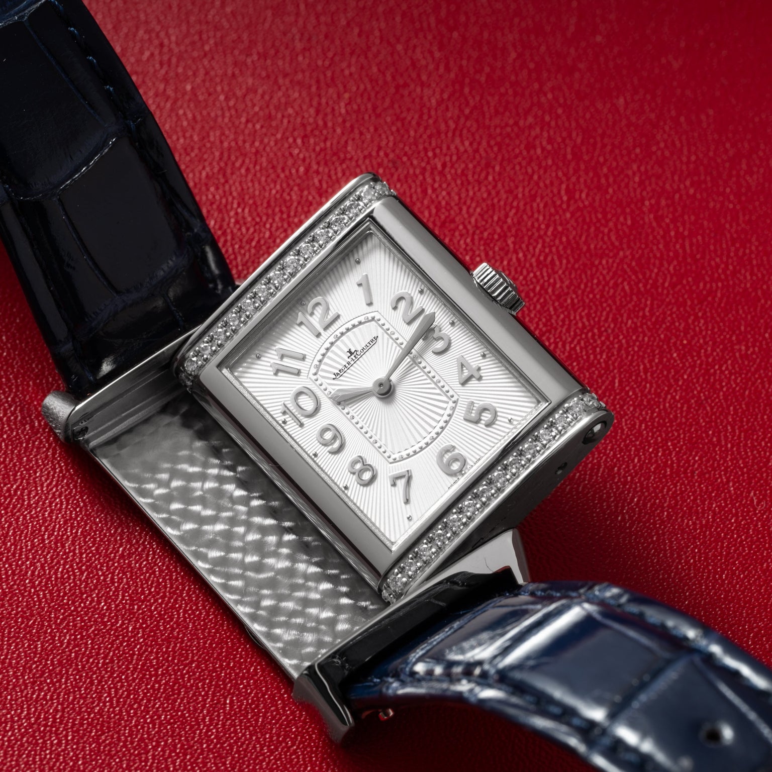 The Jaeger-LeCoultre Reverso Classique Ladies, a rectangular silver watch with a blue strap, diamond bezel, and open caseback, is displayed on a red surface.