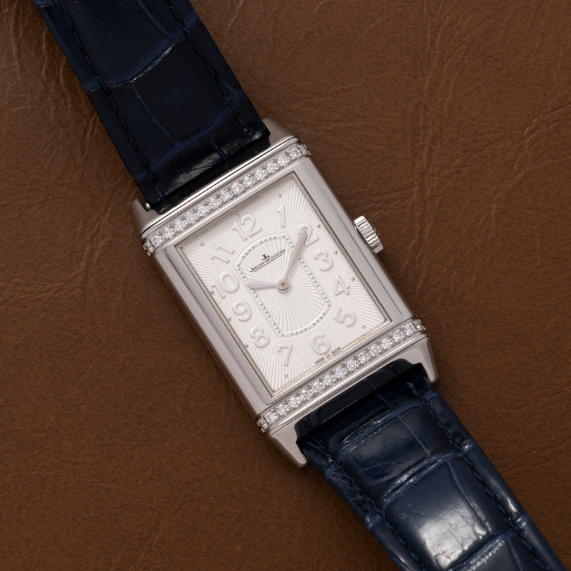 The Jaeger-LeCoultre Reverso Classique Ladies watch features a rectangular silver case, blue leather strap, and diamond accents, elegantly displayed on a brown leather surface.