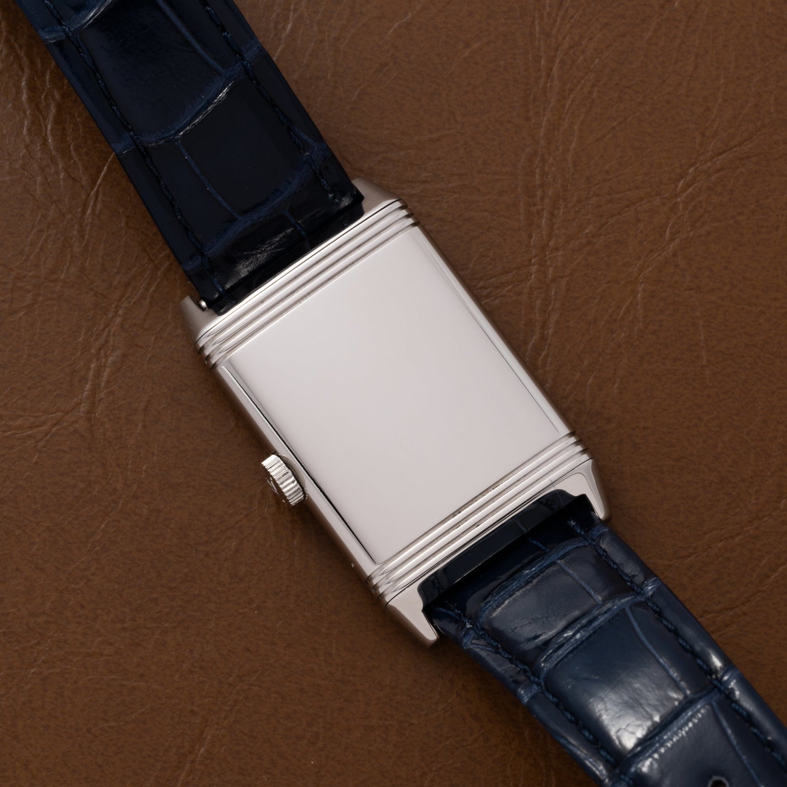 The Jaeger-LeCoultre Reverso Classique Ladies features a silver rectangular case, minimalist dial, and a blue leather strap, displayed on a brown leather surface.