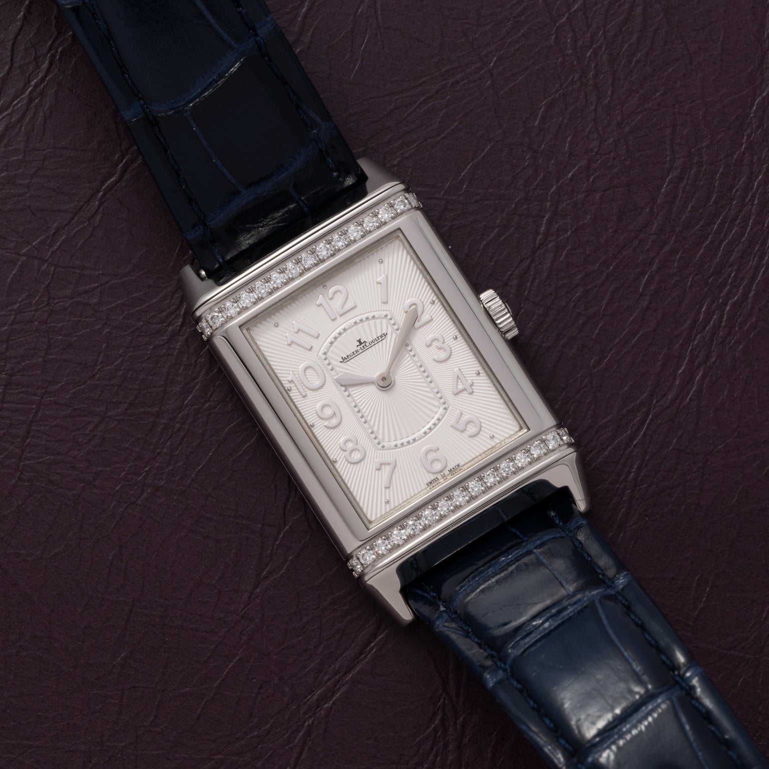 The Jaeger-LeCoultre Reverso Classique Ladies, a rectangular silver watch with diamond accents and blue leather strap, is displayed on a dark purple textured surface.